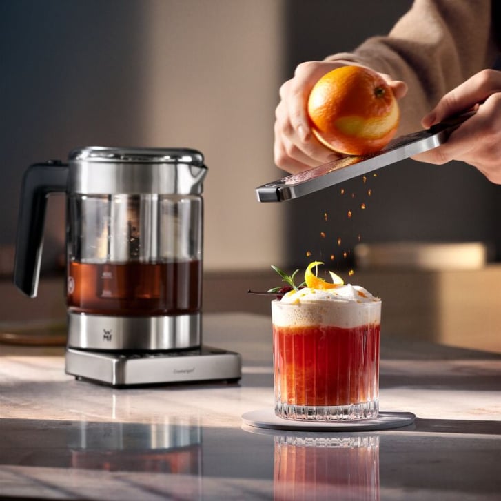 Orangenzeste wird in ein Glas gerieben, daneben steht ein Wasserkocher aus Glas